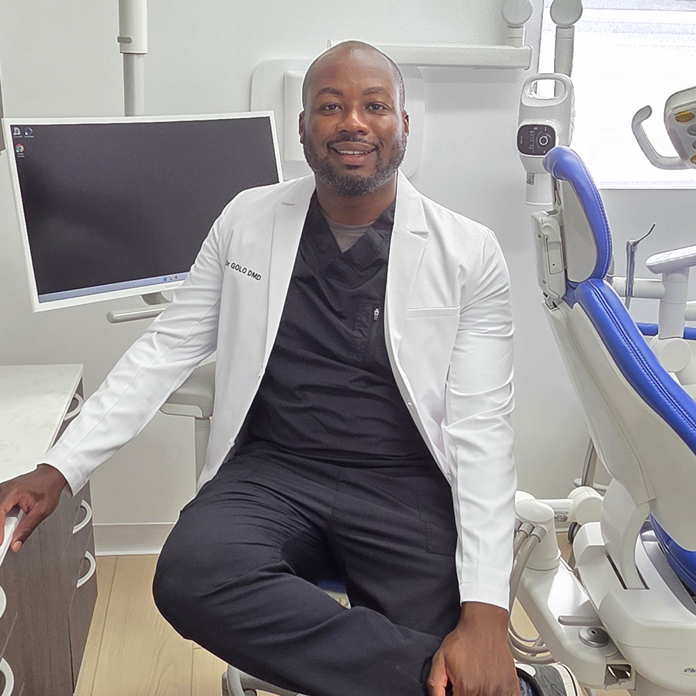 A man sitting on a dental chair, smiling at the camera, wearing a white lab coat with a stethoscope around his neck, in a dental office setting.